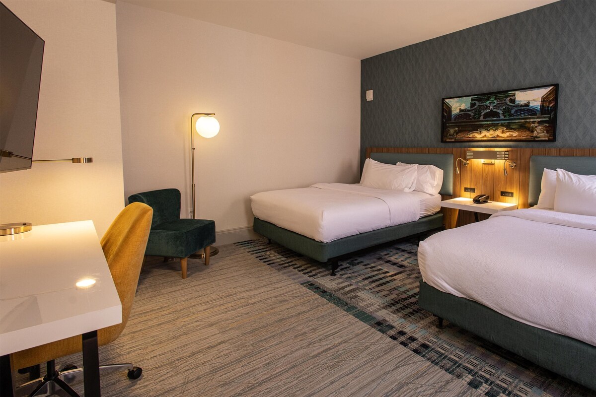 Una habitación de hotel con dos camas, un escritorio, una silla, una lámpara de pie y un cuadro en la pared.