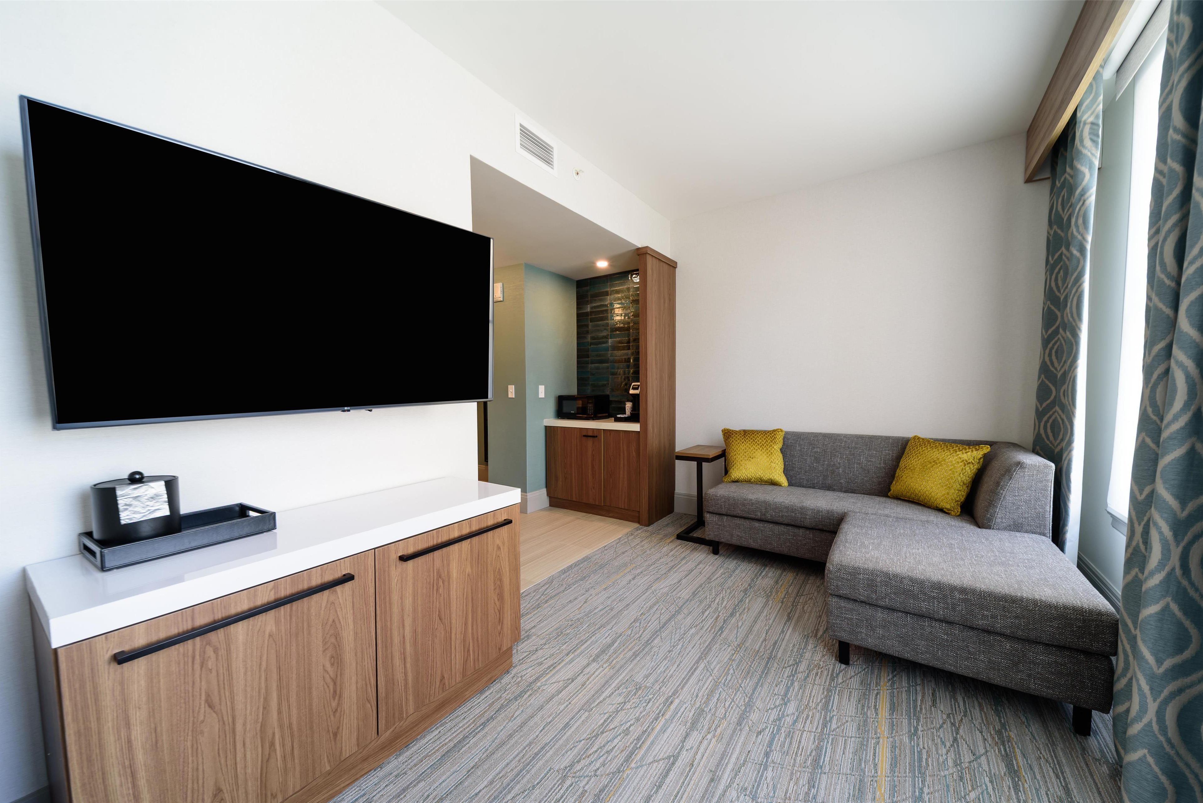 A modern living room with a flat-screen TV mounted on the wall, a grey sofa with a yellow pillow, and a wooden cabinet with a white countertop.