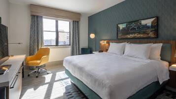A hotel room with a large bed, a desk with a chair, a window with curtains, and a painting on the wall.