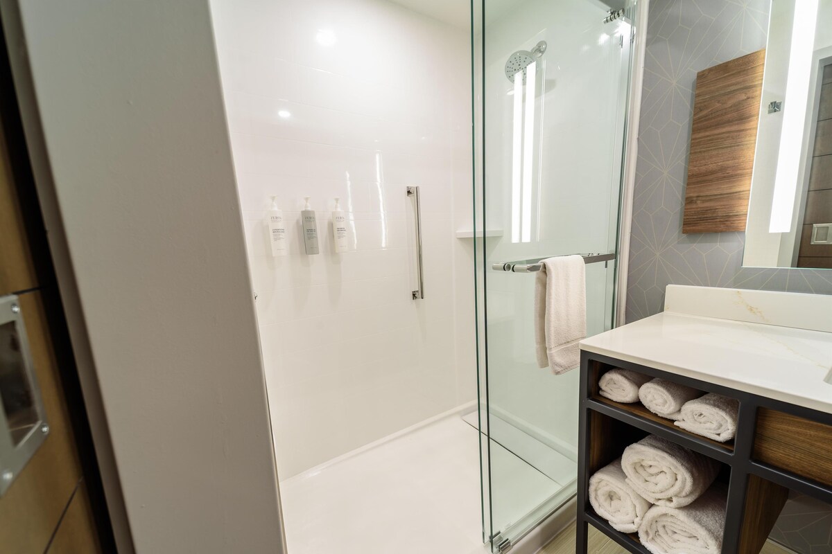 A modern bathroom with a glass-enclosed shower, a white sink, and neatly rolled towels on a shelf.