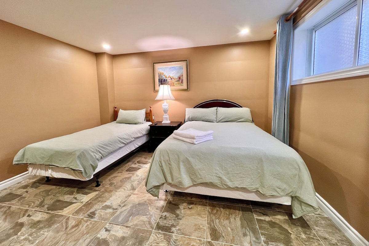 A hotel room with two single beds, a nightstand with a lamp, a painting on the wall, and a window with curtains.