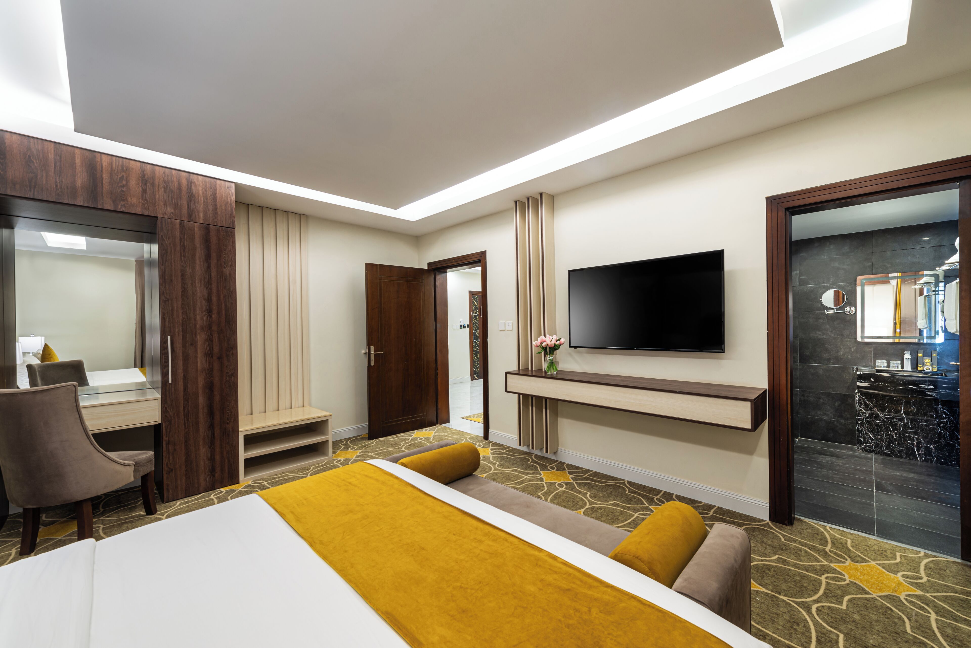 Quarto de hotel moderno com cama, escrivaninha, TV e banheiro visível através de uma porta de vidro.