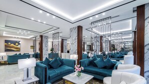 A modern hotel lobby with teal sofas, white armchairs, marble columns, and a central marble coffee table with a vase of flowers. - Garden Millennium Hotel Suites - Alsewes (Jizan)