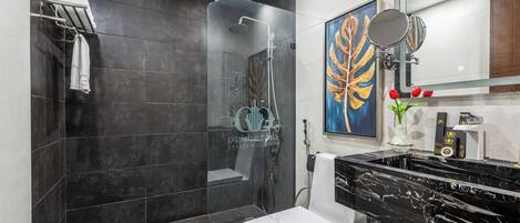 Un baño moderno con baldosas oscuras, ducha con cerramiento de vidrio, inodoro blanco e lavamanos de mármol negro.