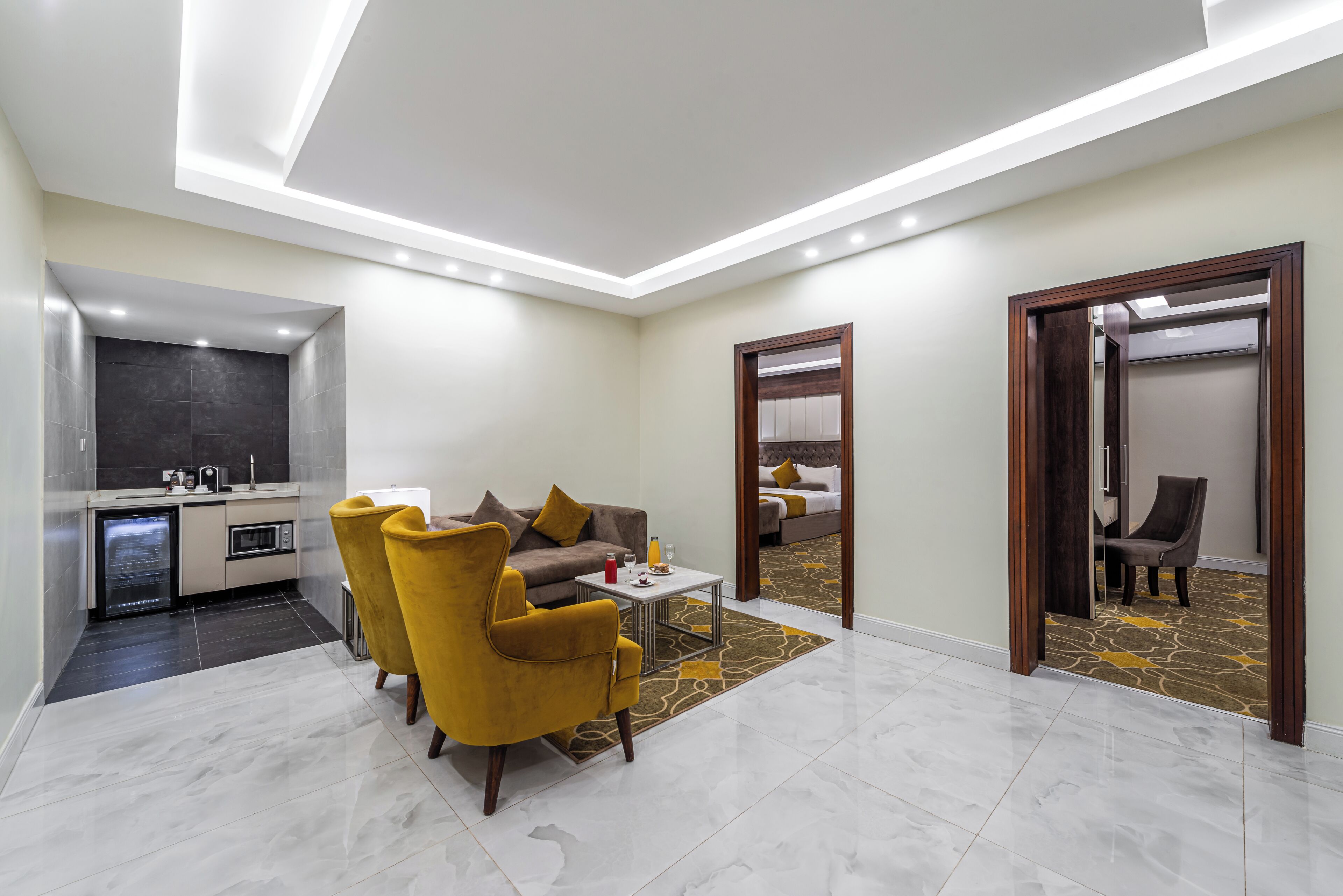 A modern hotel room with a sofa, armchairs, a small table, a kitchen area, and a bathroom.