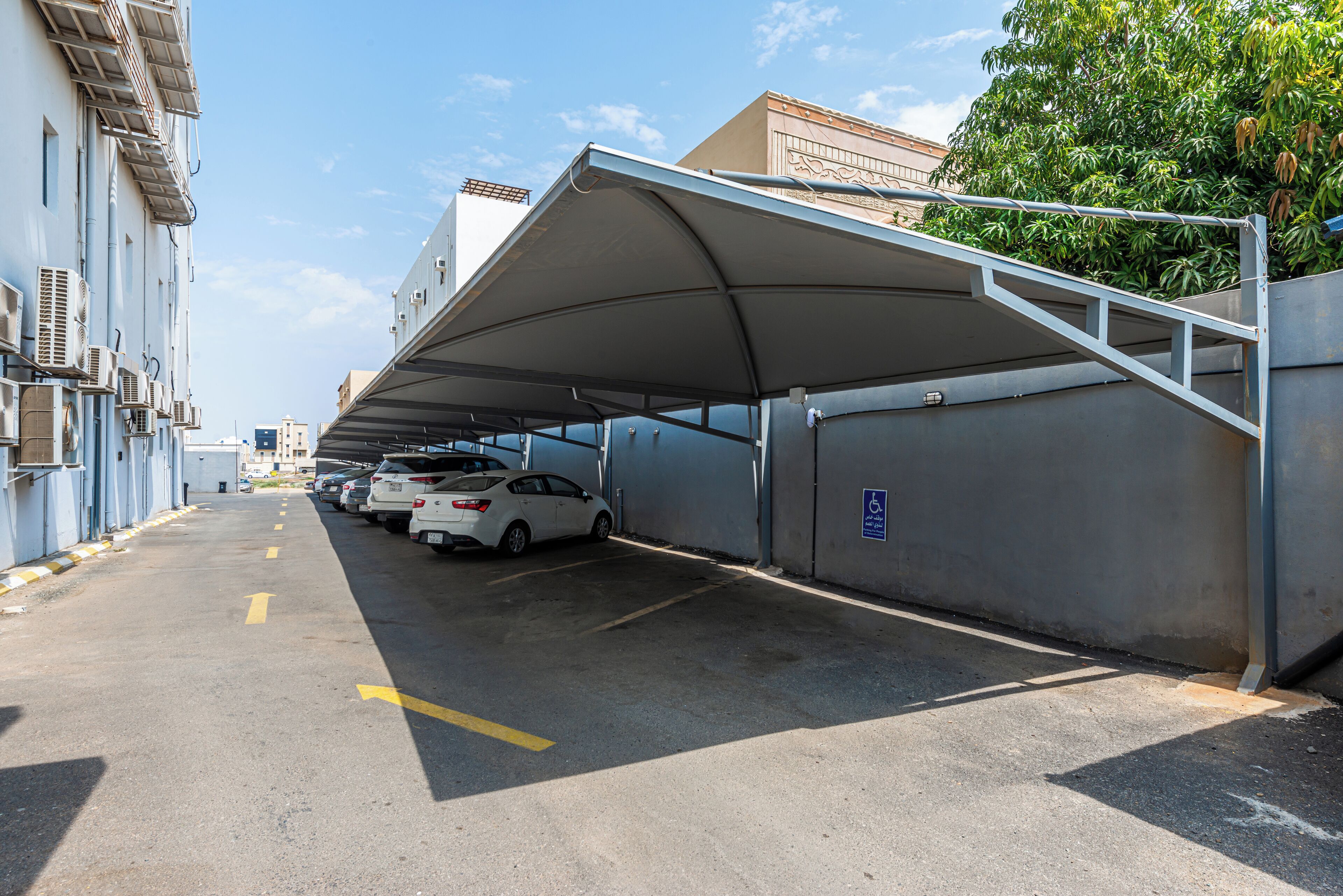 A covered parking area with cars parked under a canopy, adjacent to a building with air conditioning units.