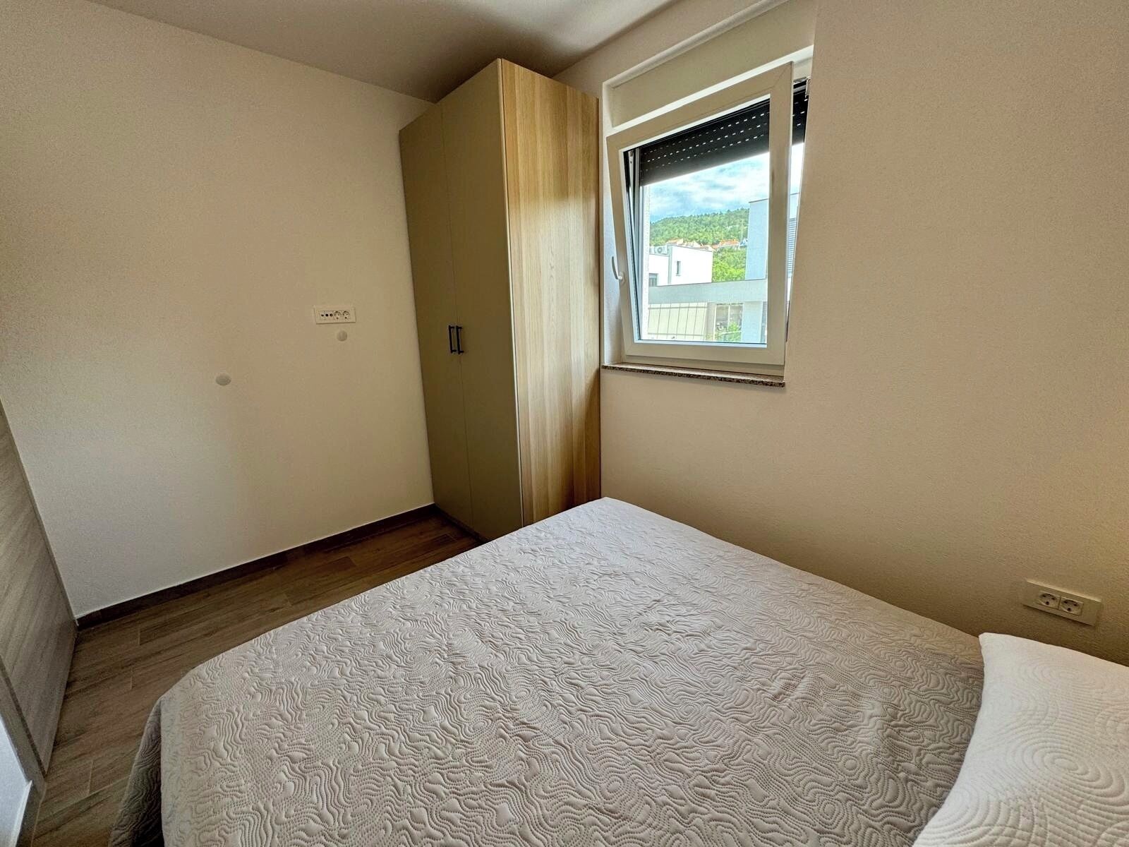 A bedroom with a bed, a wardrobe, a window, and a view of buildings outside.