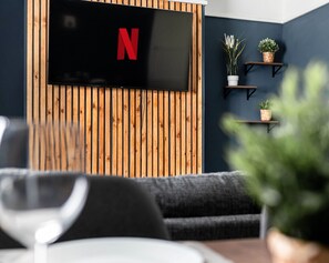 A modern living room with a flat-screen TV mounted on a wooden wall, two small shelves with potted plants, and a dark-colored sofa.