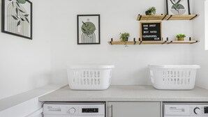 A laundry area with two washing machines, a countertop, and shelves with plants and a sign. - 4BR House | Near Sizewell (Peasenhall)