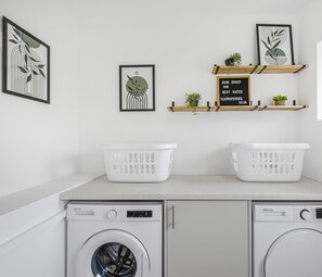 A laundry area with two washing machines, a countertop, and shelves with plants and a sign. - 4BR House | Near Sizewell (Peasenhall)