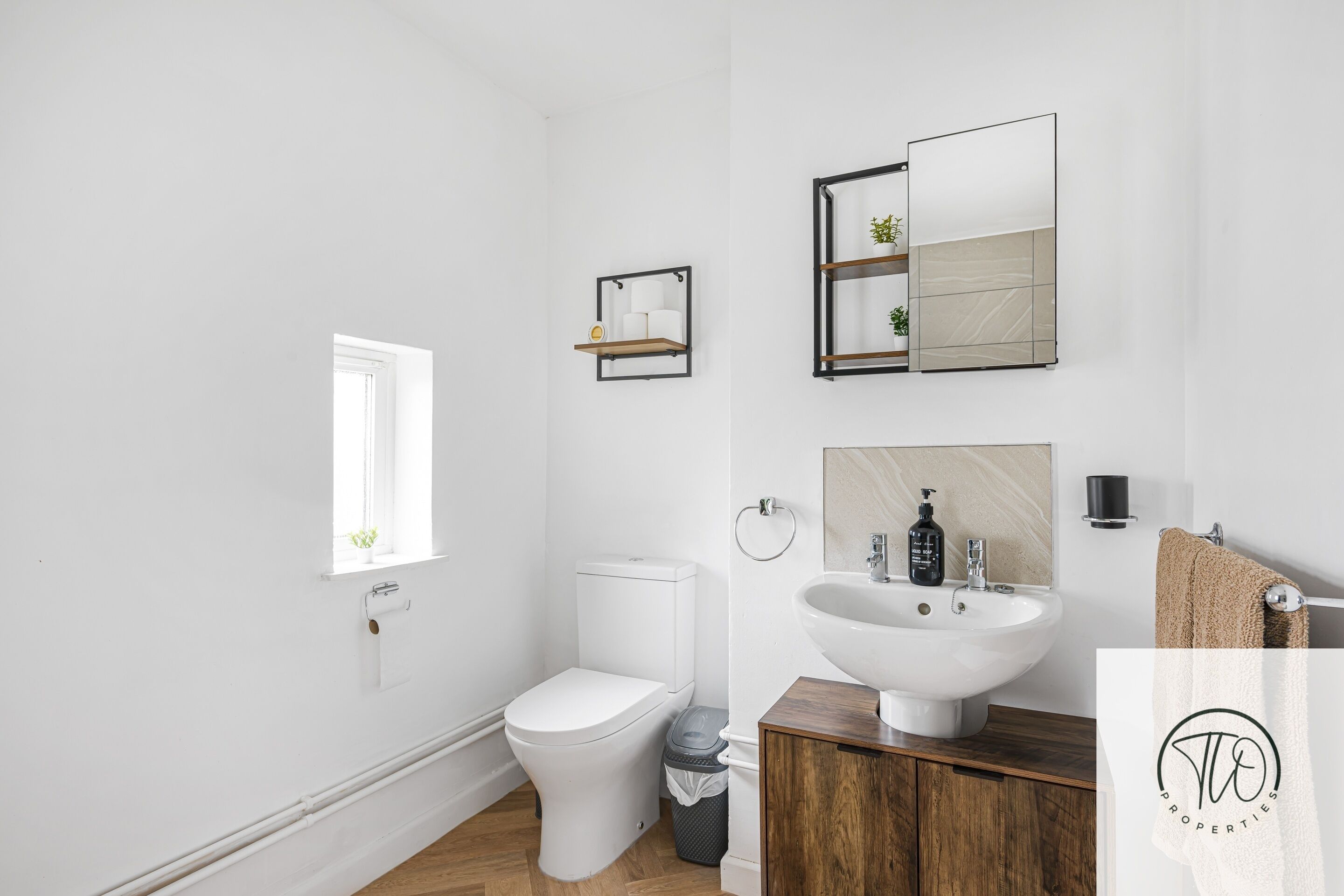 A modern bathroom with a white toilet, wooden vanity, a large mirror, and a small shelf with toiletries.
