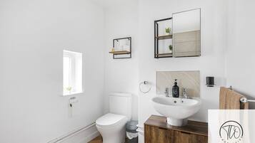 A modern bathroom with a white toilet, wooden vanity, a large mirror, and a small shelf with toiletries.