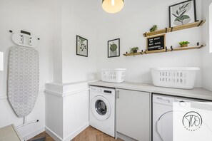 A laundry area with a washing machine, dryer, ironing board, and shelves with plants and laundry baskets.