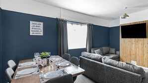 A modern dining area with a wooden table set for four, grey upholstered seating, and a large window with curtains. - 4BR House | Near Sizewell (Peasenhall)