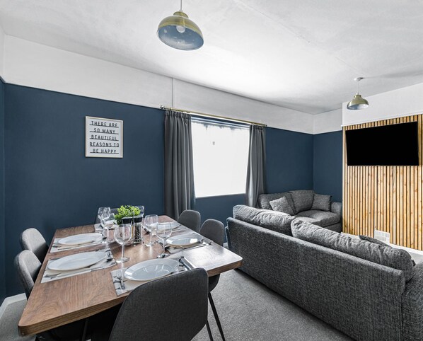 A modern dining area with a wooden table set for four, grey upholstered seating, and a large window with curtains. - 4BR House | Near Sizewell (Peasenhall)