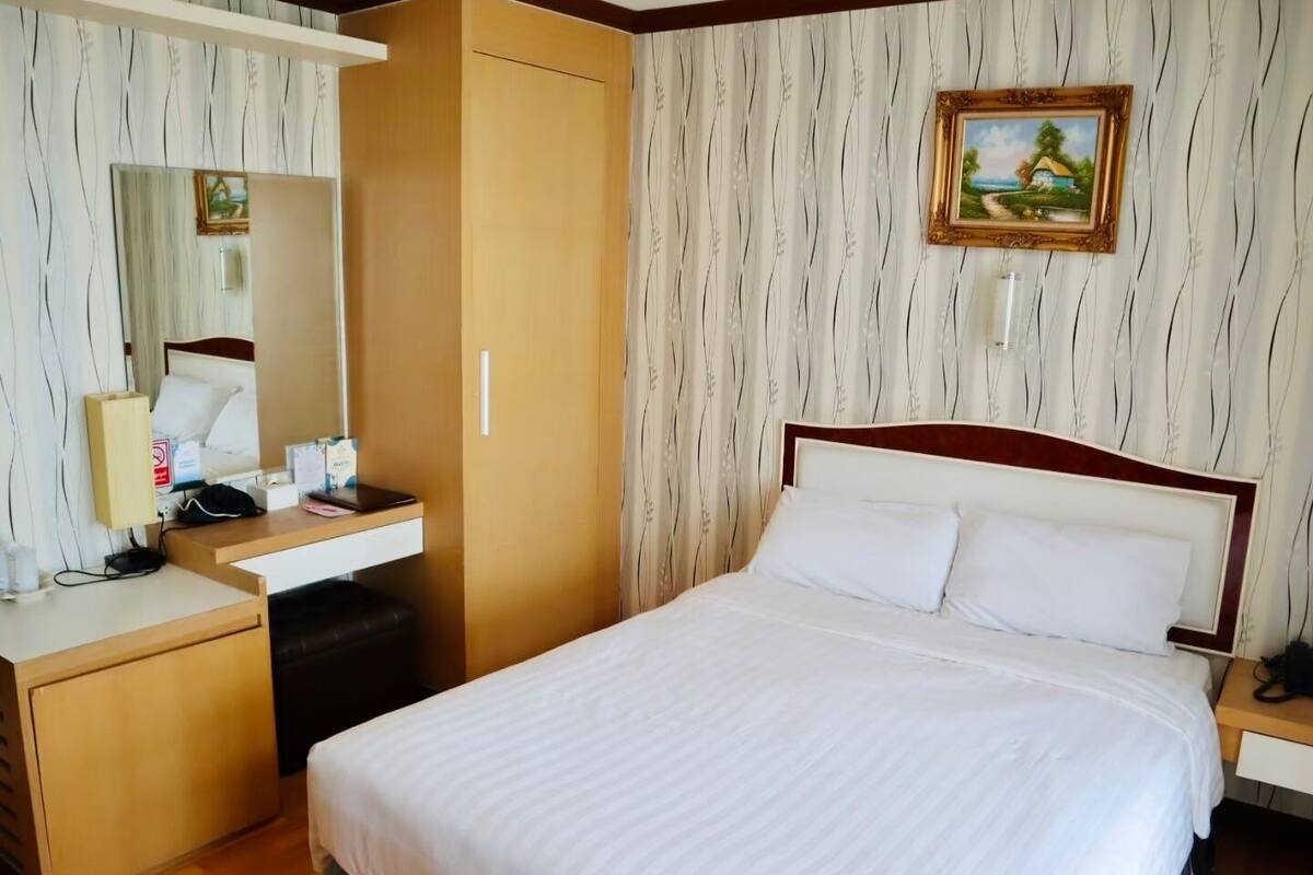 A hotel room with a bed, a desk with a chair, a wardrobe, a mirror, and a painting on the wall.