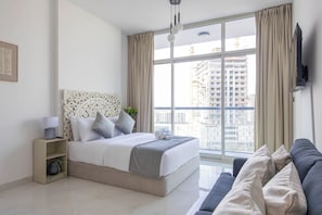 Une chambre moderne avec une grande fenêtre, un lit avec une literie grise, une table de chevet avec une lampe et un canapé gris.