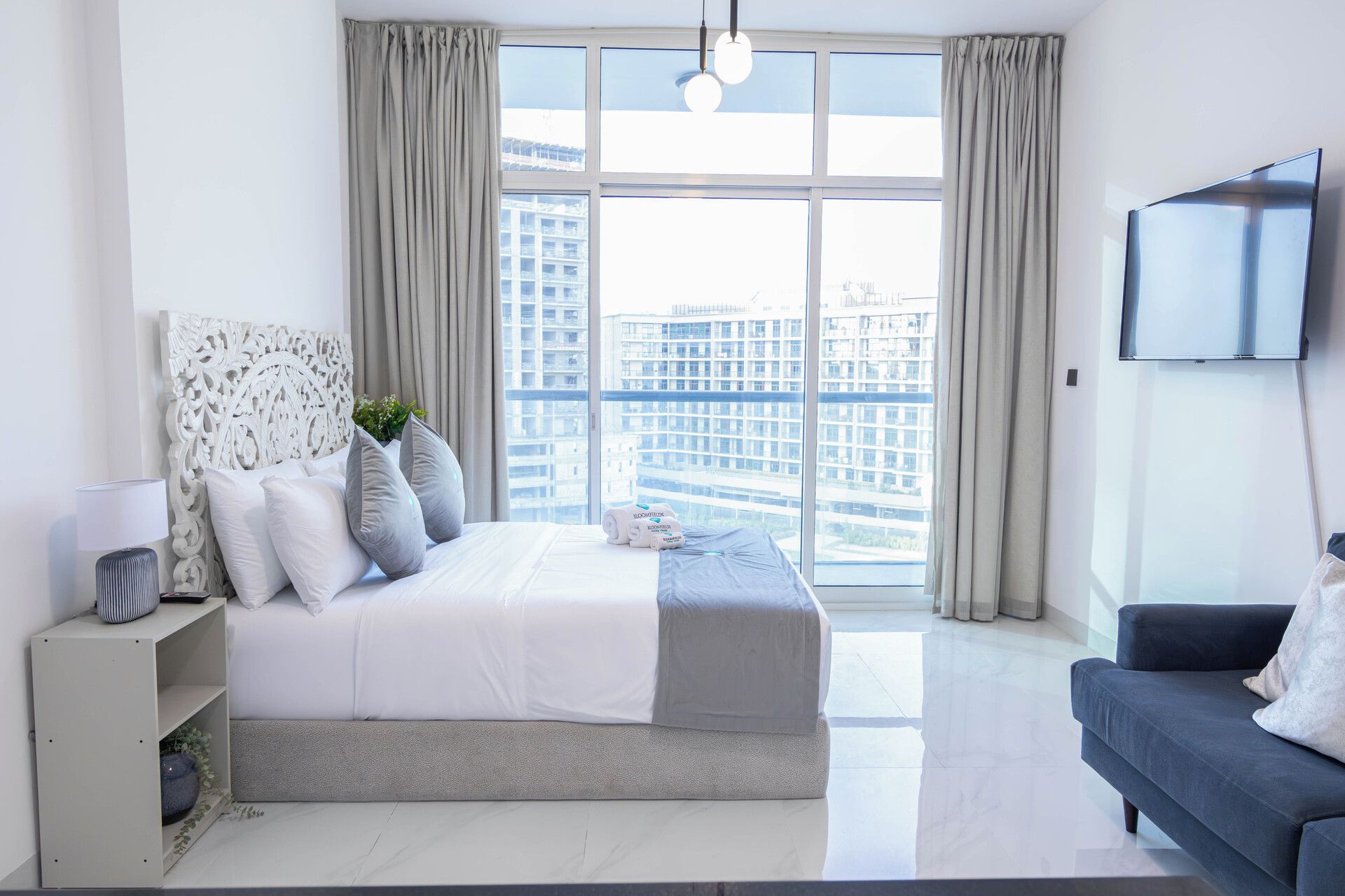 A modern bedroom with a large bed, a TV mounted on the wall, and a view of a cityscape through large windows.