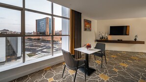 Een hotelkamer met een groot raam, een flatscreen televisie, een kleintje tafeltje met stoelen en een kamerbreed tapijt met dessin.