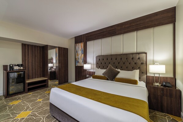 Habitación de hotel con una cama grande, cabecera de madera, minibar, escritorio y espejo.