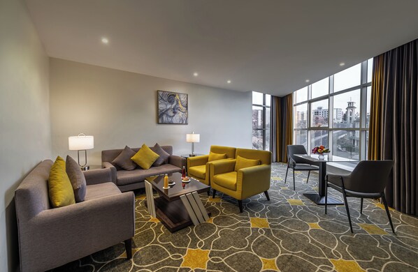 A modern hotel room with a grey sofa, yellow armchair, and a small dining table with chairs.