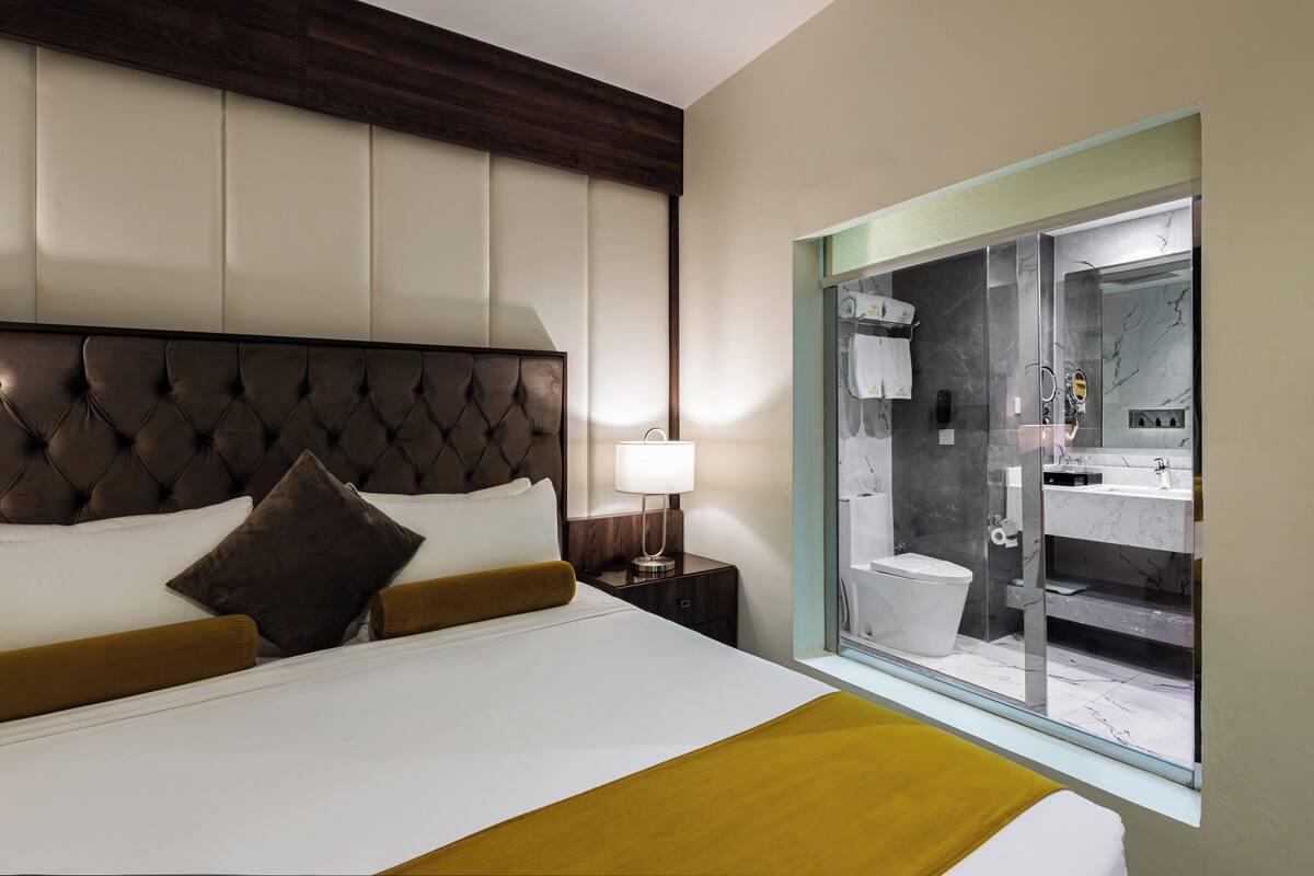 Une chambre d’hôtel avec un grand lit, une tête de lit en bois, une table de chevet avec une lampe et une vue sur la salle de bain à travers une porte en verre.