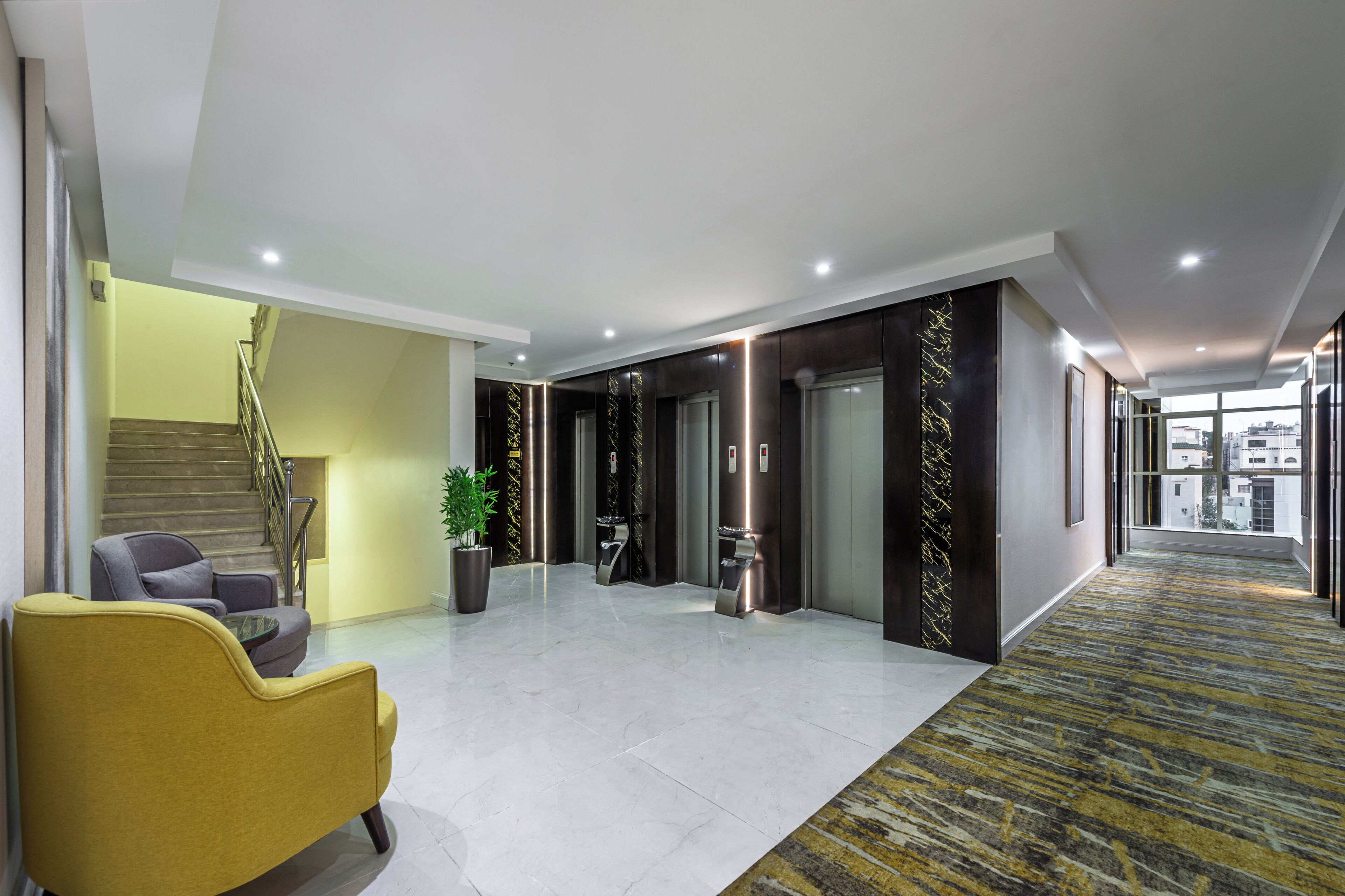 Un vestíbulo de hotel moderno con suelo de mármol, una escalera, un sillón amarillo y una planta en maceta.
