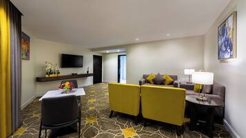 Une chambre d’hôtel comprenant un canapé, une table à manger avec un panier de fruits, un téléviseur fixé au mur et une moquette à motifs.