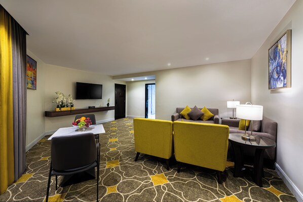 A hotel room with a sofa set, a dining table with a fruit bowl, a TV mounted on the wall, and a patterned carpet. - Garden Millennium Hotel - Abha (Abha)