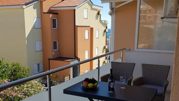An outdoor balcony with wicker furniture, a table with fruits and a bottle, and a view of neighboring buildings. - Apartments Sun Dance 2 With Heated Pool (Novalja)