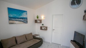 A living room with a sofa, a painting of a beach, a clock, and a door. - Apartments Sun Dance 2 With Heated Pool (Novalja)