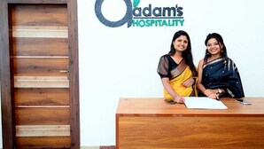 Zwei Frauen stehen an einem hölzernen Empfangstresen. Im Hintergrund ist ein Schild mit der Aufschrift 'Adam's Hospitality' zu sehen.