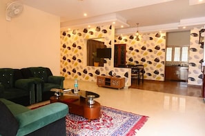 A living room with a green sofa, a coffee table, and a patterned rug. There is a TV mounted on the wall, a wooden cabinet, and a dining area with chairs. - Aadams Suites (Bengaluru)
