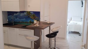 A modern kitchen with a view of a tropical beach, a dining area with two bar stools, and a connected bedroom with a white bed and nightstand. - Apartments Sun Dance With Heated Pool (Novalja)