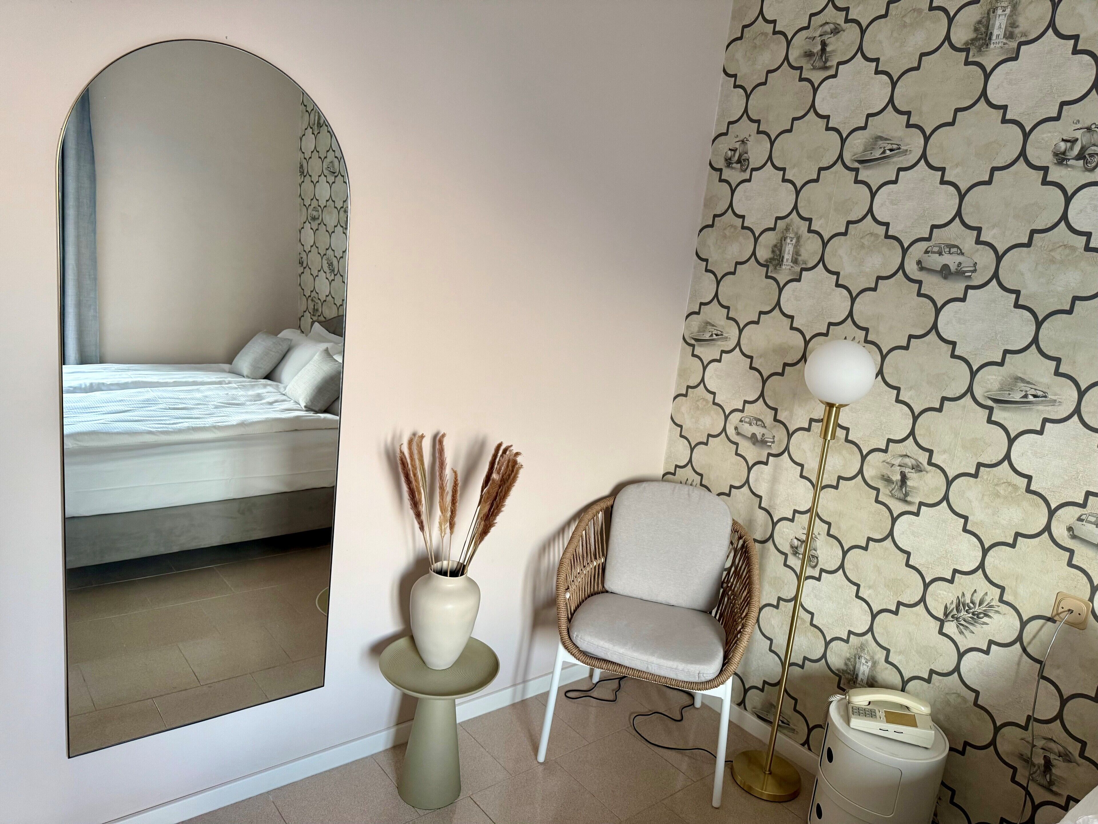 Un dormitorio con cama, espejo, jarra con plantas secas, silla de mimbre y lámpara de pie.