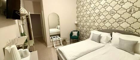 A hotel room with a large bed, a sitting area with a chair, a mirror, a flat-screen TV mounted on the wall, and a patterned wallpaper.
