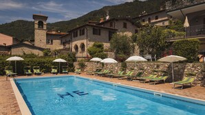 A swimming pool with lounge chairs and umbrellas, surrounded by stone buildings and a mountain backdrop. - Hotel MioMio (Tignale)