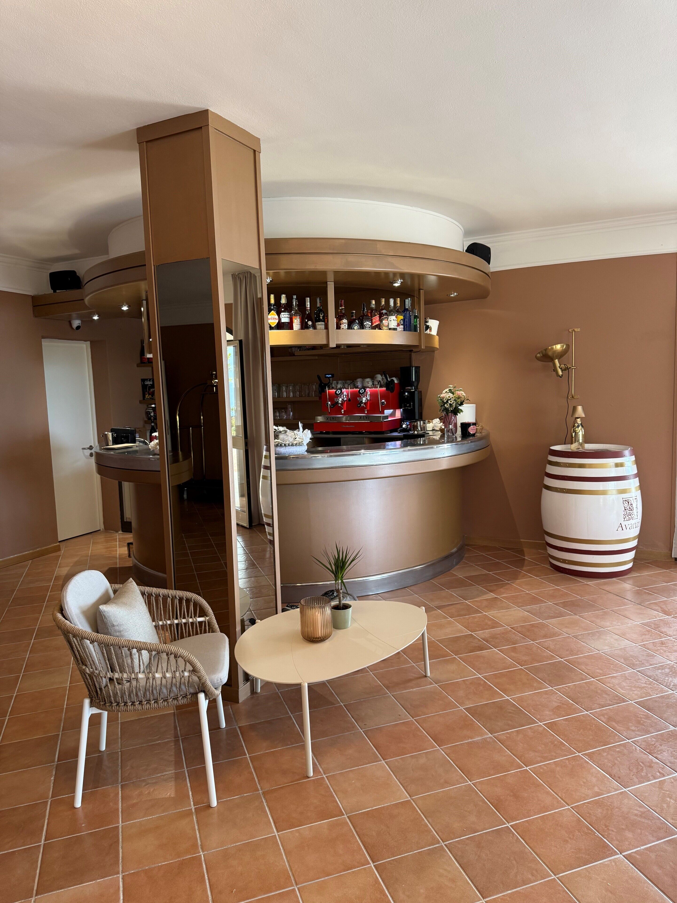 Zona bar di un hotel con una macchina da caffè espresso rossa, un tavolo rotondo con una pianta in vaso e una sedia in vimini.