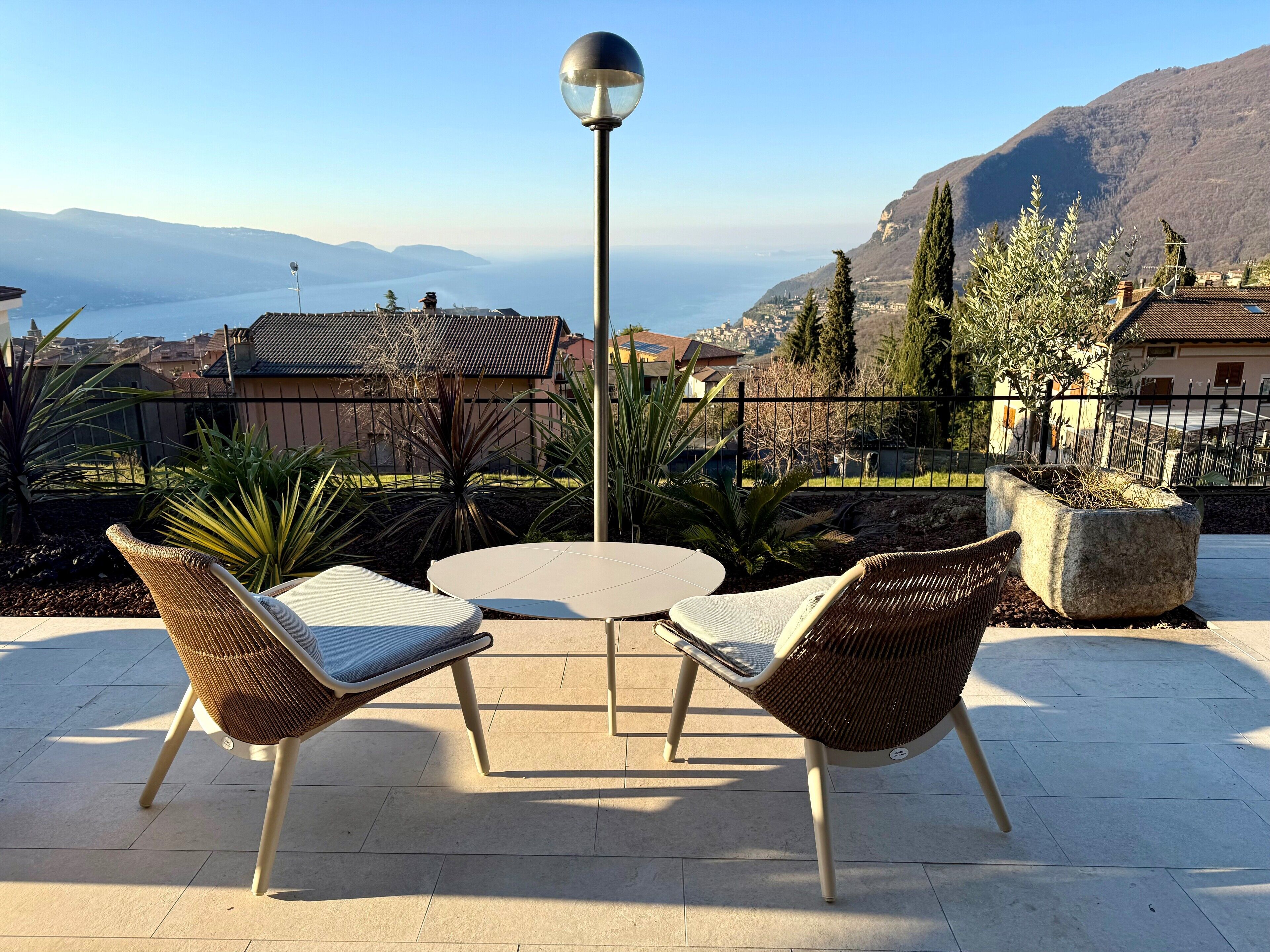 Un área de estar al aire libre con sillas de mimbre y una mesa, con vista a un paisaje montañoso y una vista despejada del mar y edificios lejanos.