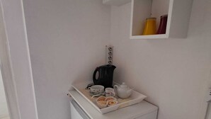 A compact kitchenette with a white refrigerator, a small countertop, and a shelf above containing a kettle and a yellow container.