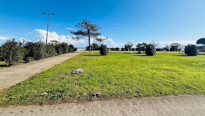 Uma trilha pavimentada serpenteia por uma área gramada bem cuidada, com árvores e arbustos, sob um céu azul claro.