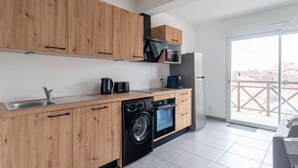 A modern kitchen with wooden cabinets, a washing machine, a refrigerator, and a window overlooking a balcony.