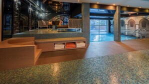 A modern indoor pool area with a mosaic floor, seating, and a view of a building through the glass wall.