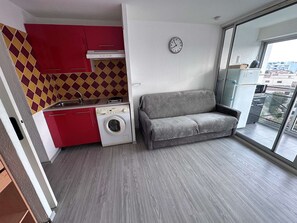 Un espacio habitable compacto con kitchenette, lavadora, sofá gris y un amplio ventanal con vistas a edificios.