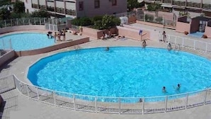 A large outdoor swimming pool surrounded by a white fence, with people swimming and lounging around.