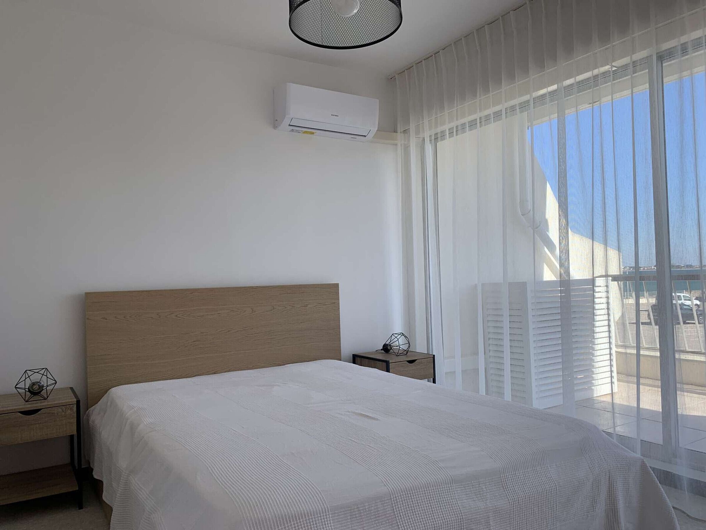 A bedroom with a wooden headboard, a white bedspread, a nightstand with a lamp, and a window with sheer curtains.