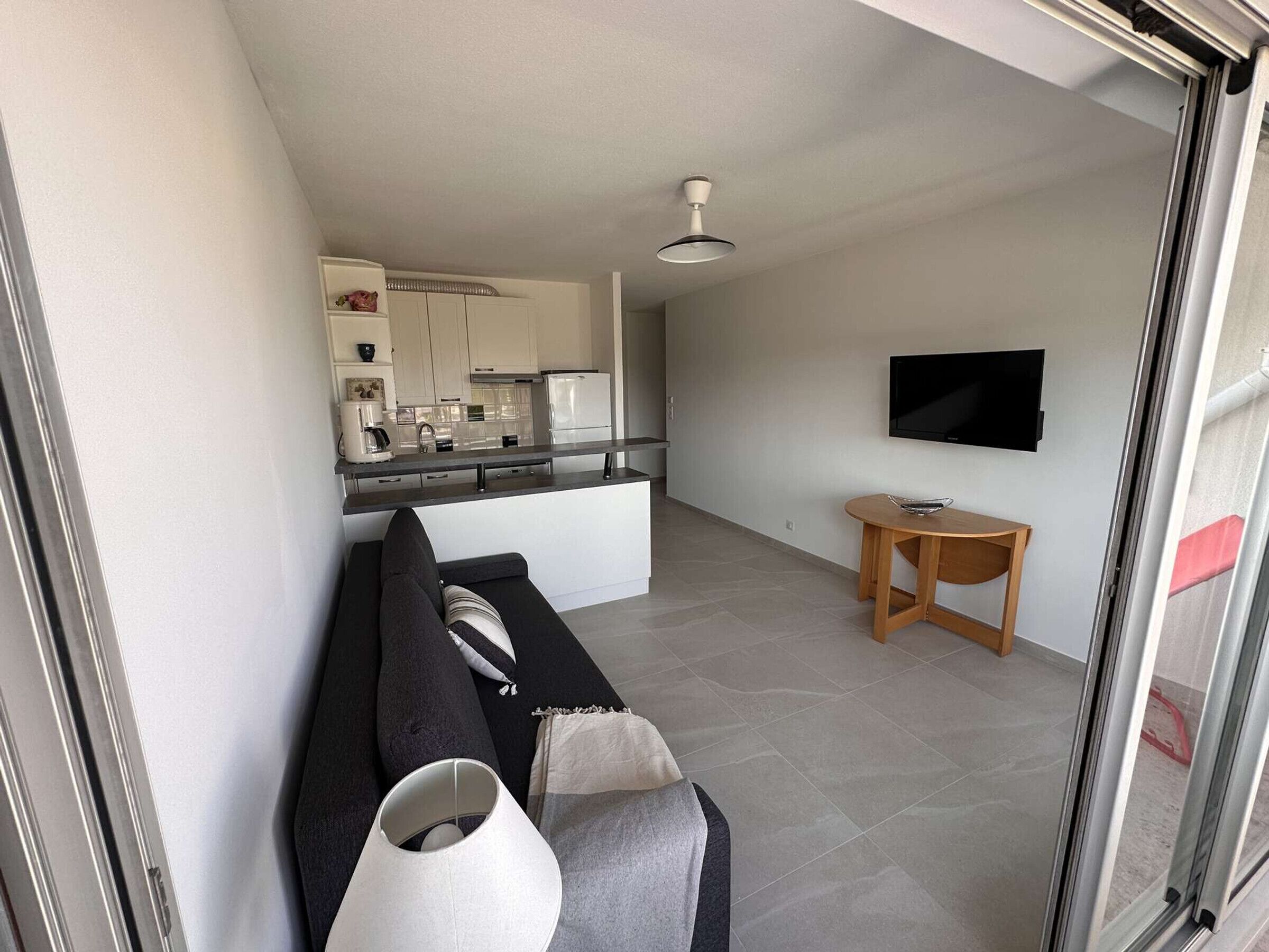 A modern living room with a sofa, a small table, a TV, and a kitchen in the background.