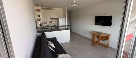 A modern living room with a sofa, a small table, a TV, and a kitchen in the background.