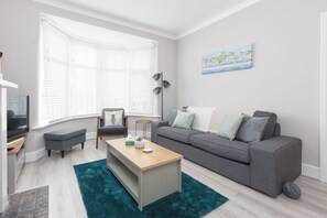 A modern living room with a grey sofa, a wooden coffee table, a teal rug, and a large window with white blinds.