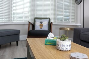 Un salon avec une table basse en bois, une boîte à mouchoirs verte, un pot blanc contenant des plantes succulentes et une chaise agrémentée d’un coussin à motif d’abeille.